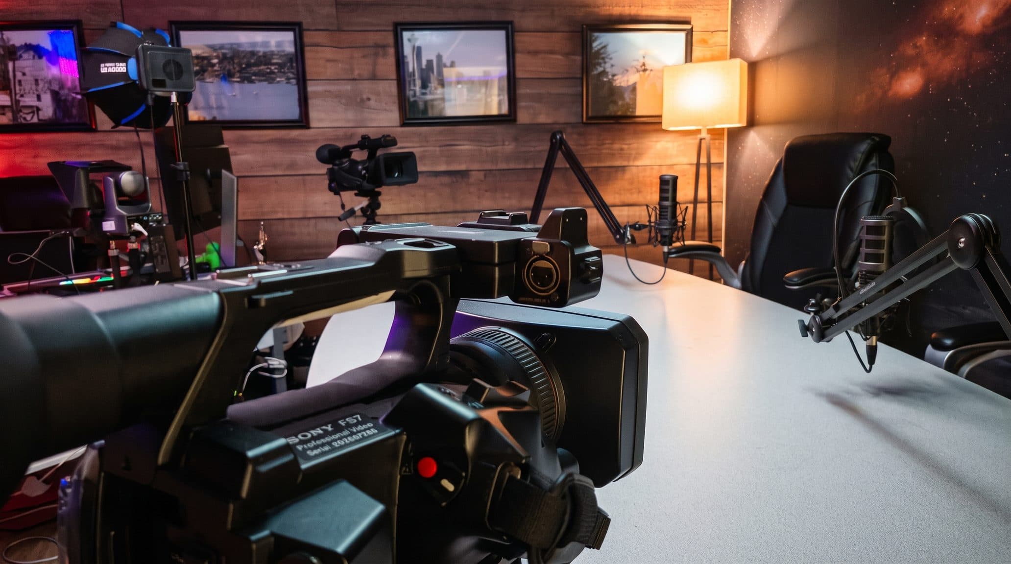 A closer view across the podcast studio table, with a professional video camera in the foreground, a second camera on a tripod and microphones on boom arms